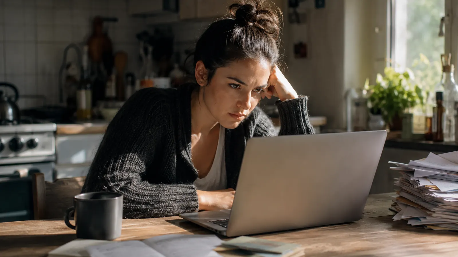 A kitchen table set up as a makeshift home office, laptop open next to a stack of mail and a coffee mug, morning light through the window