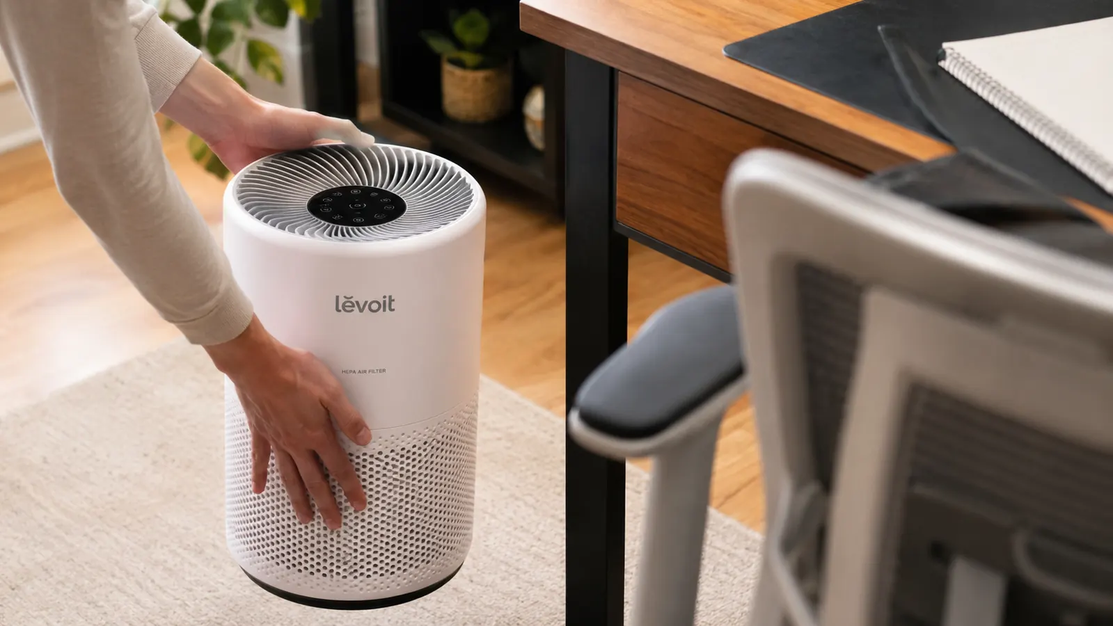 A hand setting the LEVOIT air purifier down on the floor next to a home office desk