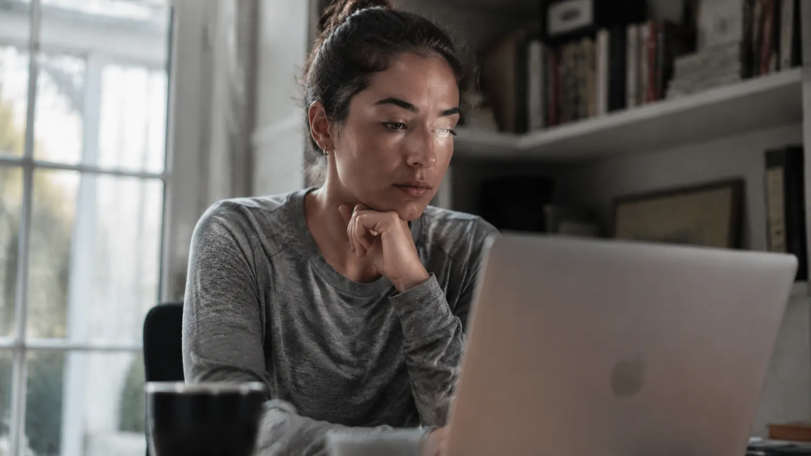 A woman on a video call at a small desk in a dim corner nook, laptop screen showing a dark, shadowed face